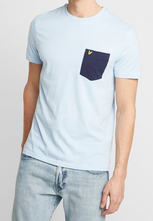 Mann trägt ein hellblaues T-Shirt mit kurzen Ärmeln, einer dunkelblauen Brusttasche und kleinem gelben Logo, kombiniert mit hellblauen Denim-Jeans.