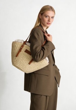 Femme blonde en tailleur marron porte un grand sac fourre-tout en paille tressée avec des sangles en cuir sur l'épaule, regardant vers la caméra.