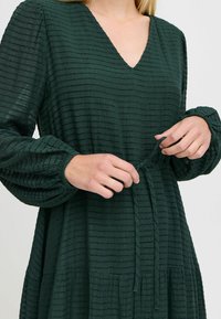 Tumma vihreä, teksturoitu mekko V-aukolla ja pallosimmeillä. Sisältää vaakasuoria raitoja ja vyön vyötäröllä, ulottuen keski-pohkeeseen.