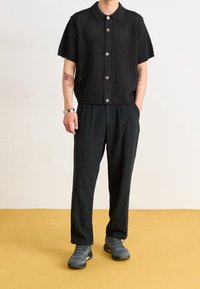 Černé pletené tričko s krátkými rukávy, límečkem a zapínáním na knoflíky, v kombinaci s volnými černými kalhotami a šedými teniskami na žluté podlaze.