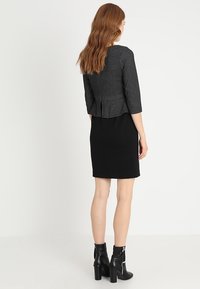 Sort kjole med en tætsiddende silhuet og en mønstret grå jakke. Outfittet har trekvartærmer og lynlåsede sorte ankelstøvler.