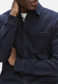 Marineblaues, strukturiertes Hemd mit durchgehender Knopfleiste, spitzem Kragen und einer einzelnen Brusttasche. Es hat lange Ärmel mit geknöpften Manschetten.