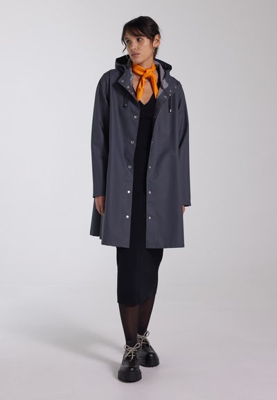 Grijze waterdichte regenjas met een capuchon, dichtgeknoopt met zilveren drukknoopjes, over een zwarte aansluitende jurk. Een oranje sjaal voegt contrast toe. Stevige zwarte schoenen.