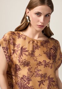 Blusa floreale bronzo con maniche corte; presenta stampe botaniche viola e un ampio scollo. La modella indossa grandi orecchini scultorei dorati.