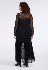 Czarna sukienka maxi z koronki z długimi rękawami, dopasowanym gorsetem, warstwową spódnicą i detalem na plecach z wycięciem, zestawiona z czarnymi botkami na kostkę.