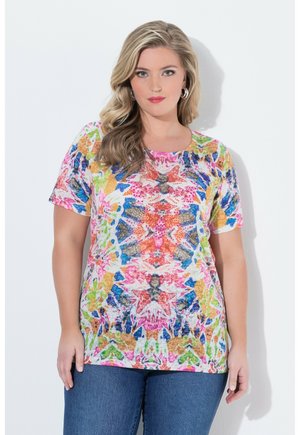 Kvinde med blondt hår iført en farverig abstrakt mønstret kortærmet top og blå jeans, står foran en ensfarvet hvid baggrund.