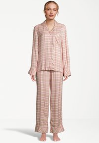 Vrouw die blootsvoets staat in een beige en roze geruite lange mouwen knoopjes pyjama top en bijpassende losse broek.
