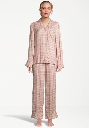 Kvinna som står barfota iförd en beige och rosa rutig långärmad pyjamasöverdel med knappar och matchande lösa byxor.