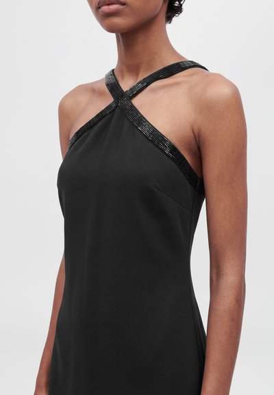 Mannequin portant une robe noire sans manches aux bretelles croisées et ornées, sur un fond clair uni.