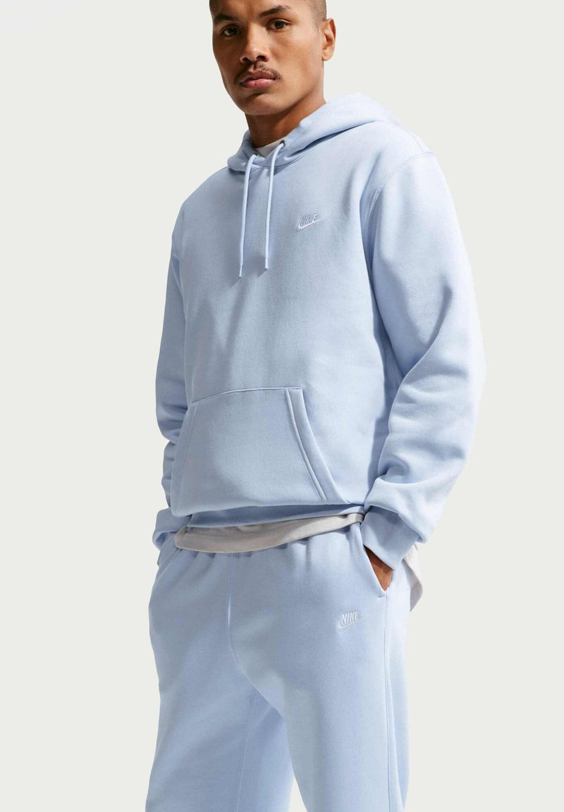 Uomo che indossa una felpa Nike azzurro chiaro e pantaloni da tuta, con le mani nelle tasche, in piedi davanti a uno sfondo semplice e chiaro.