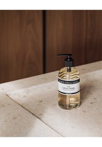 Heldere glazen fles handzeep, gelabeld "Lemon Notes," met een zwarte pompkop. Geplaatst op een gestructureerd beige aanrechtblad met gespikkelde patronen.