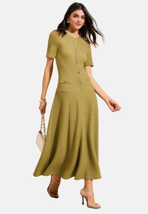 Vrouw in een textuurrijke olijfgroene midi-jurk met korte mouwen en knopen, die een beige handtas vasthoudt en beige sandalen met bandjes en hoge hakken draagt.