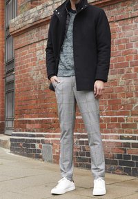 Manteau noir avec un col montant, pull gris texturé, pantalon à carreaux gris clair, et baskets blanches, le tout sur fond de mur en briques.