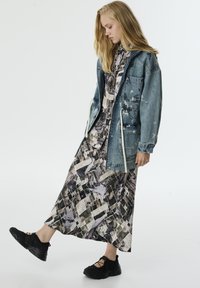 Vrouw in een geprinte maxi-jurk, oversized spijkerjack met verfspetters en zwarte sneakers, die één been iets optilt tegen een effen achtergrond.