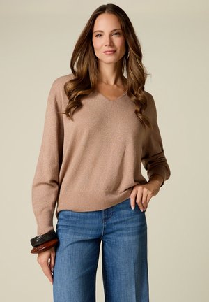 Frau mit langen braunen Haaren trägt einen beigen Pullover mit V-Ausschnitt, blaue Jeans und große Armreifen am linken Handgelenk, steht vor einem schlichten Hintergrund.