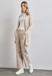 Beige cardigan över en vit skjorta, kombinerad med beige vida byxor och vita sneakers. Enkel design med fickor och knapppdetaljer.