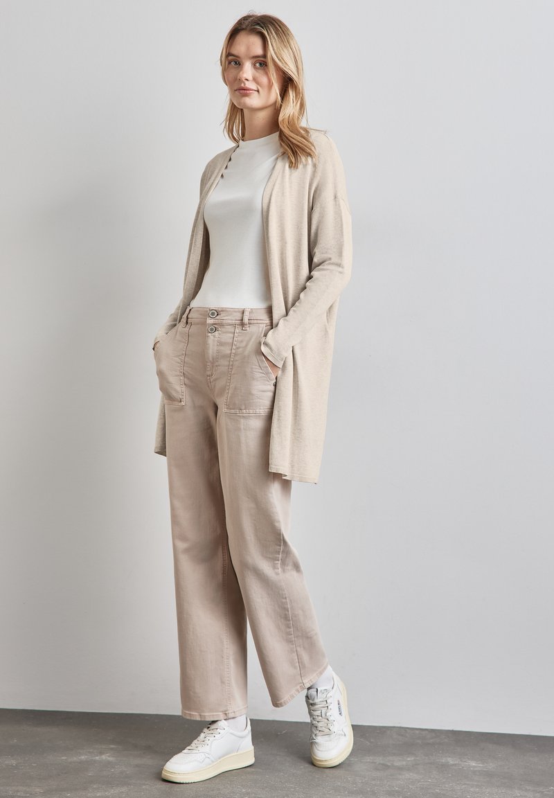 Beige cardigan över en vit skjorta, kombinerad med beige vida byxor och vita sneakers. Enkel design med fickor och knapppdetaljer.