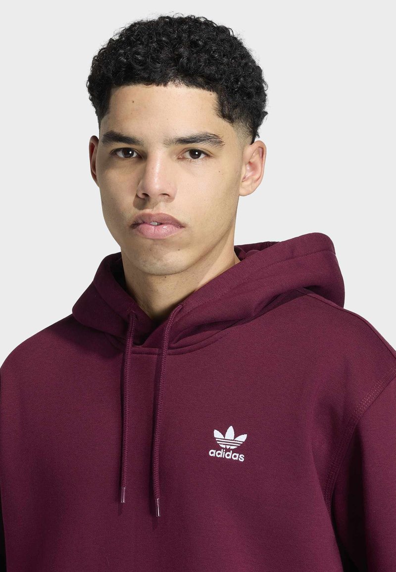 Giovane uomo con capelli corti e ricci che indossa una felpa con cappuccio Adidas color bordeaux con coulisse e logo sul petto, su sfondo neutro.