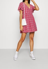 Robe florale rouge avec des manches courtes à volants et un décolleté en V, assortie à des baskets blanches et un petit sac bandoulière blanc.