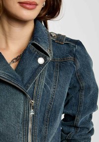 Giacca in denim con una vista ravvicinata della parte anteriore con zip, hardware argentato e cuciture a contrasto; colore blu scuro con dettagli strutturati.