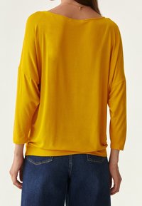 Gelbes Langarmshirt mit lockerem Schnitt, weitem Ausschnitt und überschnittenen Schultern, kombiniert mit dunklen Denim-Jeans. Glatte Textur, lässiges Design.