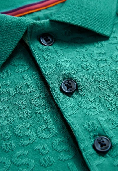 Grön polo skjorta med texturerad tyg, med ett mönster av upphöjda bokstäver, en krage och två svarta knappar. Förstärkt med färgglada ränder.