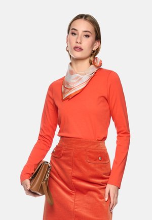 Frau trägt ein langärmeliges oranges Oberteil, einen orangen Cordrock, ein gemustertes Halstuch und hält eine braune Handtasche vor einem einfarbigen Hintergrund.