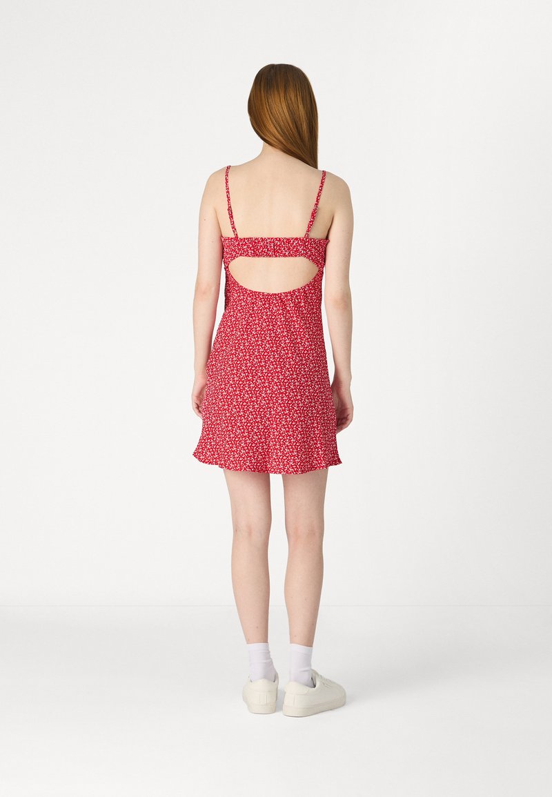 Rotes Blumenkleid mit Spaghettiträgern, offenem Rücken, ausgestelltem Saum und leichtem Stoff. Trägt weiße Sneaker und Knöchelsocken.