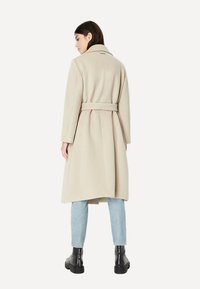 Beige Wollmischungsmantel mit tailliertem Gürtel, langen Ärmeln und einem Schlitz hinten, kombiniert mit hellblauen Jeans und schwarzen Stiefeletten.