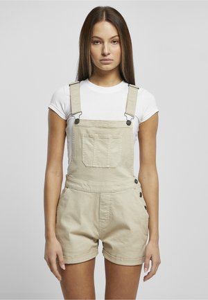 Femme aux longs cheveux raides portant une chemise blanche à manches courtes et une salopette courte beige, debout devant un fond gris uni.
