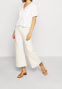 Chemise blanche à manches courtes boutonnée avec manches bouffantes, associée à un pantalon large crème et des sandales marron à talons blocs à lanières.