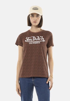 Femme portant une casquette beige Von Dutch, un t-shirt Von Dutch California à imprimé léopard marron et un jean bleu, debout devant un fond blanc.