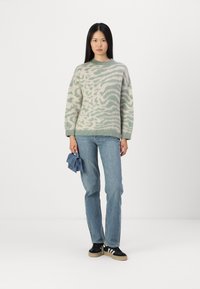 Maglione verde chiaro con una texture pelosa, caratterizzato da un motivo ondulato; abbinato a jeans blu e sneaker nere, mentre tiene una piccola pochette blu.