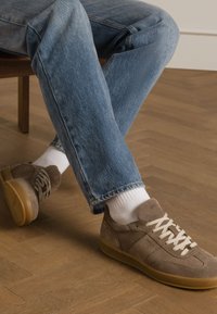Braune Wildleder-Sneaker mit einer glatten, leichten Sohle, beige Schnürsenkeln, strukturierten Akzenten und kombiniert mit hochgekrempelter heller Denimhose und weißen Socken.