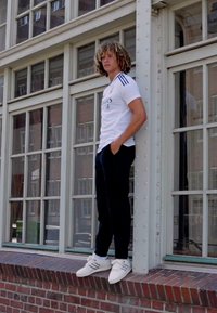 T-shirt sportivo bianco con strisce nere, pantaloni neri e sneaker bianche. Persona in piedi su un ledge di mattoni accanto a grandi finestre.