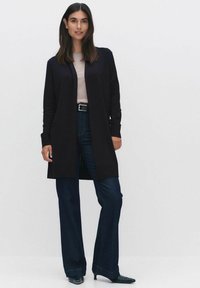 Zwarte lange cardigan met open voorkant, gecombineerd met een lichtbeige top en donkere spijkerbroek met flare, met een aansluitende taille en stijlvolle zwarte schoenen.
