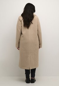 Beige tekstureret lang frakke med lige snit, enkelt rygslids og to sidelommer, parret med mørke jeans og sorte sko.