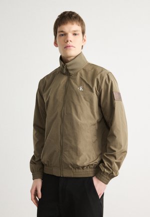 Giacca kaki con colletto alto, zip completa, polsini elastici e orlo. Presenta una toppa con logo in rilievo sulla manica. Tessuto liscio e leggero.