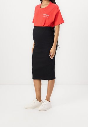 Gravid person iført en rød "MAMA" t-shirt, sort figursyet midi-nederdel og hvide sneakers, stående mod en ensfarvet hvid baggrund.