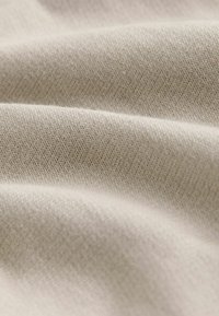 Beiger Stoff mit einer glatten Oberfläche, der feine diagonale Rippungen aufweist und in sanften Falten drapiert ist, wodurch ein nahtloses Erscheinungsbild entsteht.