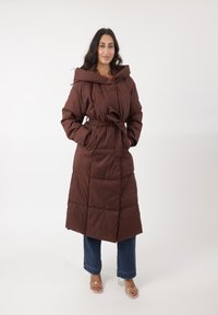 Bruine pufferjas met een tailleband, oversized capuchon en gequilt textuur. Gekleed over blauwe jeans en doorzichtige hakken sandalen.