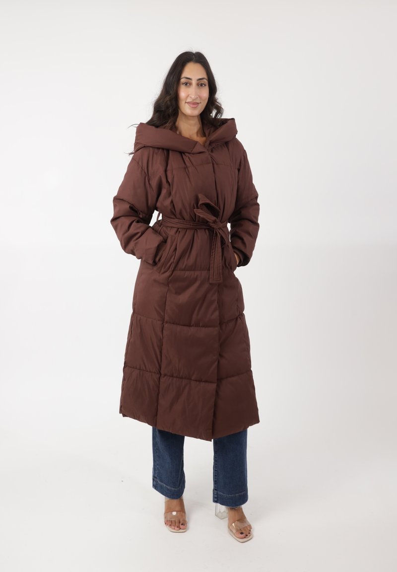 Abrigo puffer marrón con cintura ceñida, capucha extragrande y textura acolchada. Llevado sobre jeans azules y sandalias de tacón transparente.