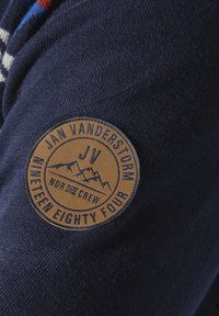 Navy-Blau gestrickter Stoff mit einem runden, braunen Leder-Patch, auf dem die relieffierte Schrift "JAN VANDERSTORM", "JV" und "NEUNZEHNHUNDERT VIER" zu sehen ist.
