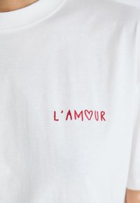 Bijela majica s crvenim izvezenim tekstom "L'AM♡UR" na prsima, s simbolom srca koji zamjenjuje slovo "O".