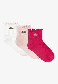 Kolm paari soonikkoes sokke: valged, heleroosad ja erkroosad. Igal paaril on volditud ülaosa ja tikitud roheline krokodilli logo.