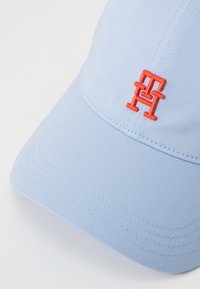 Căciulă de culoare albastru deschis, realizată dintr-un material în dungi, cu un logo portocaliu brodat. Designul include o margine curbată și detalii cusute.