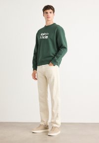Felpa verde con logo bianco "Calvin Klein", pantaloni beige a gamba dritta e sneakers beige. Design semplice, texture morbida, vestibilità rilassata.