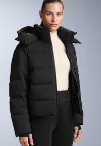 Crna zimska jakna s podstavom i visokim ovratnikom, sa prednjim zatvaračem i gumbima na pritisak. Nosena preko kreme jakne s rebrastim uzorkom i prilagođenim crnim hlačama.