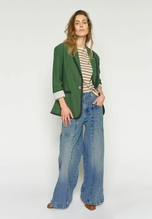 Frau trägt weit geschnittene Patchwork-Jeans, gestreiftes Hemd und grünen Blazer mit hochgekrempelten Ärmeln, posiert vor einem schlichten weißen Hintergrund.