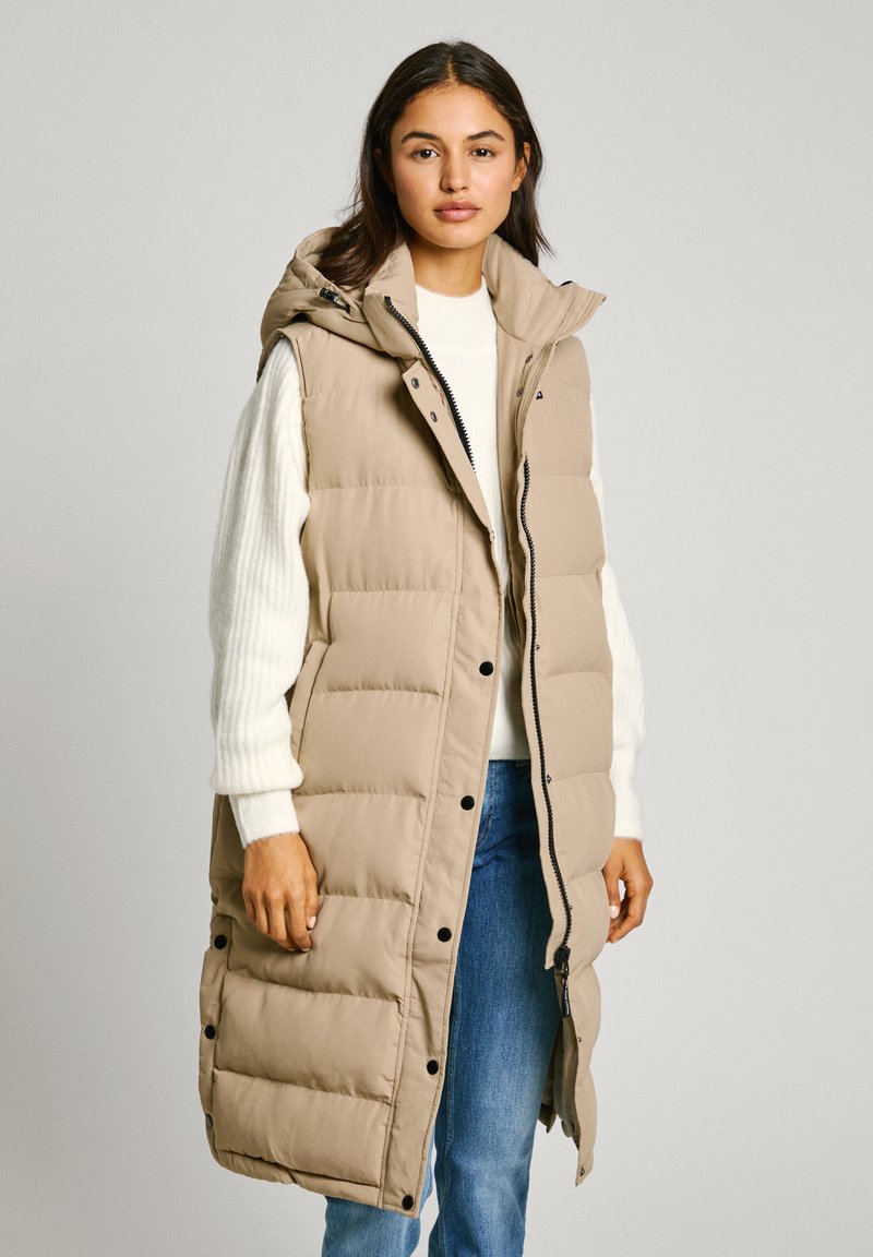 Beige lång puffer väst med huva, som har dragkedja och tryckknappsstängning, sidofickor och ett quiltat mönster. Bärs över en vit tröja.